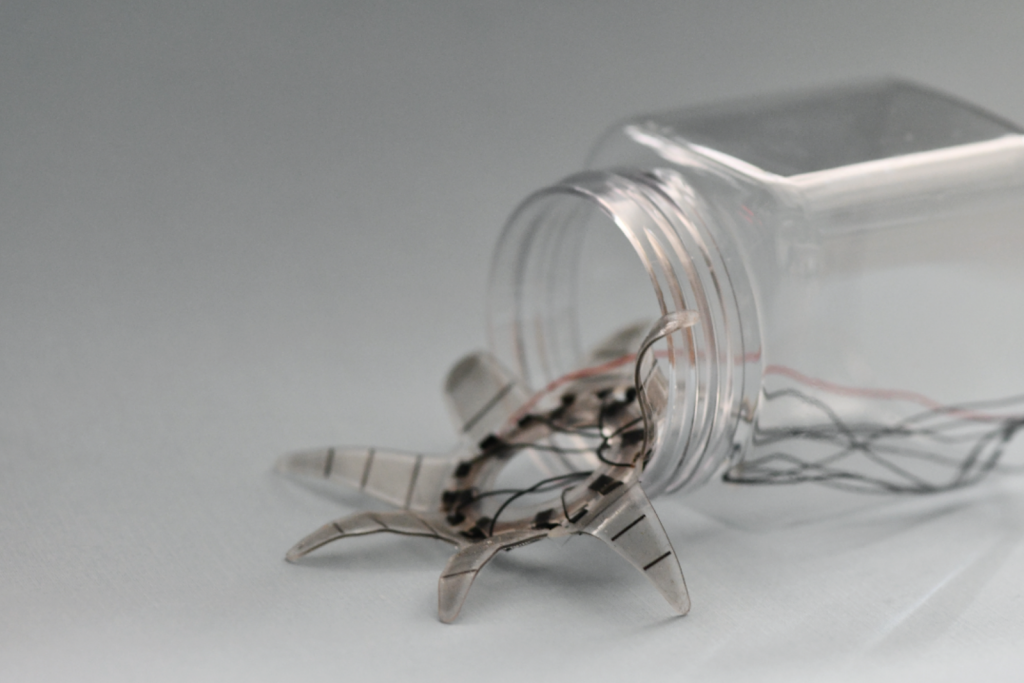 Clear plastic jar laying on its side with a small, translucent, flexible 8-legged soft robot crawling out with several wire leads trailing behind.